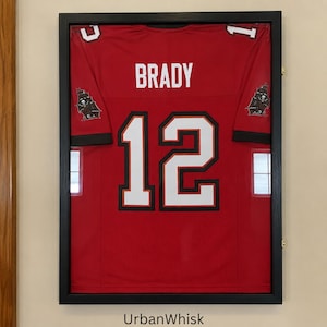 May include: A red sports jersey with the name BRADY and the number 12 in white with black outlines, displayed inside a rectangular black frame. The jersey features small pirate ship logos on each sleeve and is encased behind a clear glass panel.