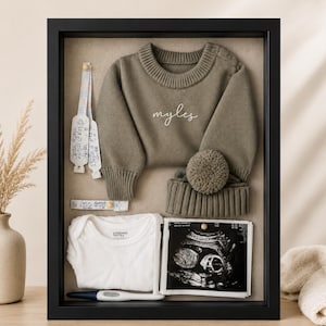 May include: A black shadow box frame containing a sage green knit sweater with the name myles embroidered in white, a matching pom-pom beanie, a white bodysuit, a digital thermometer, two hospital identification bands, and an ultrasound scan.
