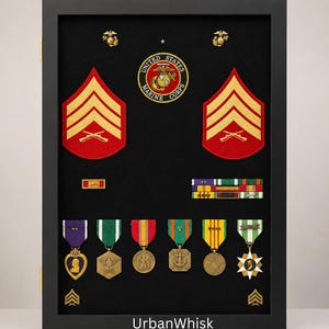May include: A black rectangular display case containing United States Marine Corps memorabilia, including two red and gold sergeant chevrons, a circular emblem, various colorful military ribbon bars, and six hanging medals on a black background.