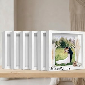 May include: A set of six white square shadow box frames arranged in a row. The front-most frame contains a photograph of a couple embracing in a field, decorated with dried pampas grass and cotton stems. The text UrbanWhisk is printed on the frame.