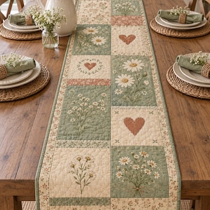 May include: A quilted table runner featuring a patchwork design of sage green and cream squares. The pattern includes embroidered white daisy clusters, small floral prints, and solid heart shapes in a muted terracotta colour, all stitched with a grid texture.