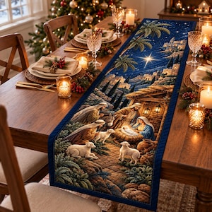 May include: A rectangular fabric table runner featuring a quilted nativity scene with a dark blue starry sky, palm trees, and stone buildings. The central illustration depicts figures gathered around a manger with sheep in a landscape.