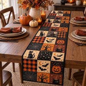 Op de afbeelding: Een rechthoekige gewatteerde tafelloper met een patchworkontwerp van vierkanten met een Halloween-thema. Het patroon bevat zwarte katten, oranje pompoenen, vleermuizen, spinnenwebben, ketels en ruitjesblokken in zwart, oranje en crème.