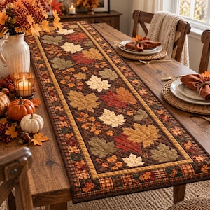 May include: A rectangular quilted table runner featuring a pattern of autumn maple leaves in shades of red, orange, yellow, and green on a dark brown background, bordered by a plaid fabric strip with small leaf motifs along the edges.
