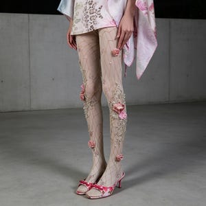 Könnte beinhalten: Beige, transparente Strumpfhose mit zartem Netzmuster, verziert mit dreidimensionalen rosa Stoffrosen, weißen Kunstperlen und filigraner Silberstickerei. Die Strumpfhose wird mit rosa, floral gemusterten, offenen Sandaletten kombiniert.