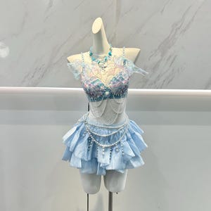 Puede incluir: Un conjunto de dos piezas azul claro que incluye un top tipo sujetador con adornos iridiscentes en forma de escamas y mangas con volantes, combinado con una minifalda a juego de volantes escalonados. Adornado con cadenas de perlas blancas.