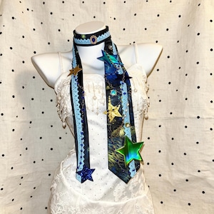 Puede incluir: Un conjunto de gargantilla y corbata larga con estampado de noche estrellada, ribete de encaje azul claro y una gema ovalada azul central. El conjunto está decorado con varios dijes metálicos y brillantes en forma de estrella y cadenas de cuentas azules.
