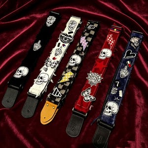 May include: Five guitar straps arranged side-by-side, featuring various fabric textures and colors including black, white, checkered, red, and blue denim. Each strap is decorated with assorted embroidered patches, including skulls, skeletons, butterflies, lightning bolts, and hearts.