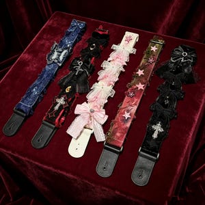 May include: Five guitar straps arranged side-by-side. The first is blue lace with butterfly appliqués. The second is black with red accents, a cross, and bows. The third is pink with layered bows. The fourth is red with star patches. The fifth is black with bows and a cross.