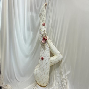 May include: Off-white fishnet tights featuring a diamond pattern and embellished with decorative fabric roses in shades of pink and red, along with intricate gold-toned lace appliqué detailing along the side of the leg.