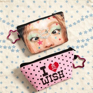 May include: Two zippered pouches with star-shaped carabiners. The top pouch features a close-up face print with the text "GIRLS WHO THRIFT" and colorful stars. The bottom pink pouch has black polka dots and the text "I WISH" with a red heart.