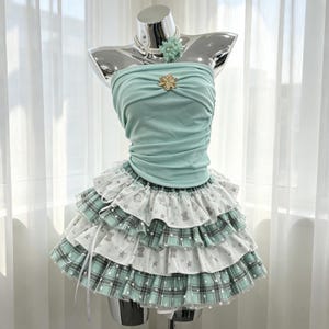 May include: A mint green strapless tube top with a gold star-shaped embellishment and a matching mint green floral choker. The skirt is a multi-layered, ruffled design featuring alternating tiers of white fabric with grey star prints and mint green plaid.