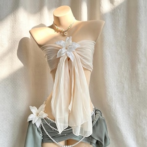 May include: A beige strapless tube top featuring a sheer, flowing fabric overlay gathered at the centre with a large white flower. The garment is paired with a sage green skirt adorned with a white floral accent and a layered pearl chain belt.
