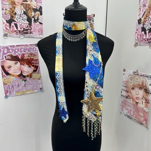 May include: A blue and yellow fabric choker and scarf set featuring white lace trim, a central amber-colored gem, silver cross charms, and blue glitter star appliqués. The scarf includes dangling star-shaped charms and beaded fringe accents.
