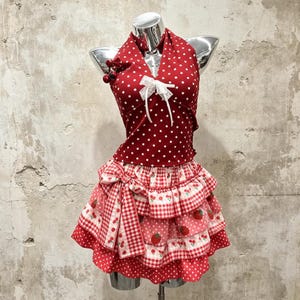 Puede incluir: Un top halter rojo con lunares blancos y un lazo de encaje blanco, combinado con una falda de capas con estampado de cuadros vichy rojo y blanco, fresas y lunares rojos, decorada con un lazo lateral grande y dijes de cerezas.