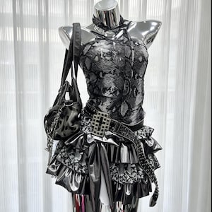 Könnte beinhalten: Ein ärmelloses Oberteil mit schwarz-weißem Schlangenmuster und passendem Halstuch sowie ein Stufenrock aus metallisch silbernem und Schlangenmuster-Stoff. Eine schwarze Handtasche mit Silberketten und ein schwarzer Gürtel mit Kreuzschnalle sind dabei.