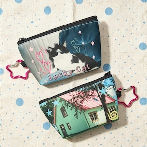 May include: Two zippered pouches with black trim and pink star-shaped carabiners. The top pouch features a black and white cat wearing sunglasses with the text Lucky Cat and pink hearts. The bottom pouch displays a green house with a pink roof.