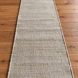 May include: A long, rectangular runner rug featuring a woven texture with a blend of natural tan, beige, and off-white fibers. The rug has a consistent, tight weave pattern throughout its entire length and is finished with clean, straight edges.