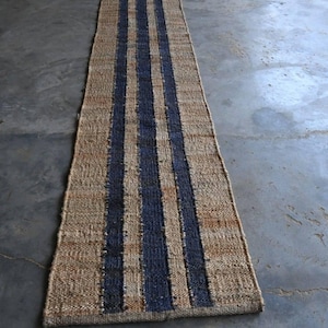 May include: A long, rectangular runner rug woven from natural tan fibers, featuring three distinct vertical stripes in a deep navy blue color. The texture of the rug is coarse and braided, with a consistent weave pattern running the entire length.