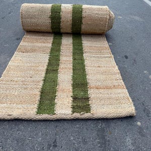 May include: A rectangular woven runner rug made of natural tan fibres featuring two parallel dark green stripes running vertically down the centre. The rug is partially unrolled, showing a textured, coarse surface with a consistent weave pattern.