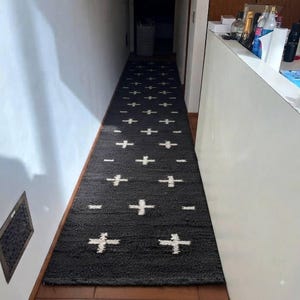 Peut inclure: Un long tapis de couloir rectangulaire présentant une couleur de base gris anthracite foncé avec un motif répétitif de petits symboles blancs en forme de croix disposés en grille sur toute la surface du revêtement de sol.