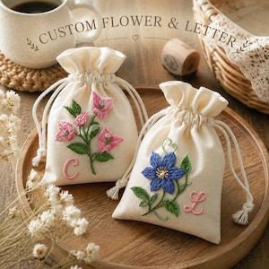 Könnte beinhalten: Zwei cremefarbene Stoffbeutel mit Kordelzug und individueller Stickerei. Ein Beutel zeigt eine rosa Blume und den Buchstaben C, der andere eine blaue Blume und den Buchstaben L. Der Text CUSTOM FLOWER & LETTER ist über den Beuteln zu sehen.