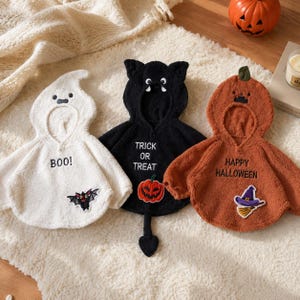 May include: Three hooded capes in white, black, and orange fleece. The white cape features a ghost hood, the text BOO!, and a bat patch. The black cape has a cat hood, the text TRICK OR TREAT, and a pumpkin patch. The orange cape has a pumpkin hood, the text HAPPY HALLOWEEN, and a witch hat patch.