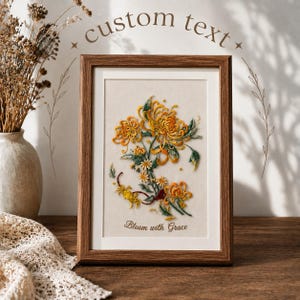 Puede incluir: Un marco de madera rectangular que contiene una pieza bordada sobre tela de color blanco roto. El bordado representa flores de crisantemo amarillas y naranjas con hojas y tallos verdes. El texto "Bloom with Grace" está bordado en marrón.