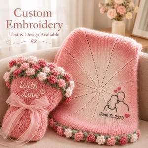 May include: A pink knitted blanket and a matching pink knitted accessory adorned with crocheted flowers. The blanket features an embroidered outline of a couple, two red hearts, and the text June 12, 2023. The accessory includes a sheer ribbon.