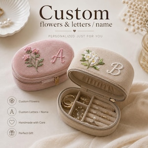 Puede incluir: Dos joyeros ovalados de terciopelo, uno rosa empolvado con flores rosas bordadas y la letra A, y otro beige con flores blancas bordadas y la letra B. El joyero beige está abierto, mostrando compartimentos interiores con varios anillos.