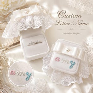 May include: Two white velvet ring boxes featuring lace trim and a satin bow with a heart charm. One box is open, displaying a silver ring, while the other is closed with the embroidered name "amy" in pink, white, and blue thread on the lid.