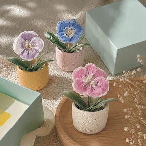 Puede incluir: Tres pequeñas flores artificiales en maceta con pétalos de tela bordados en tonos lavanda, azul y rosa. Cada flor está colocada en una maceta de cerámica texturizada y moteada con el texto "elf planet" grabado en la base frontal.
