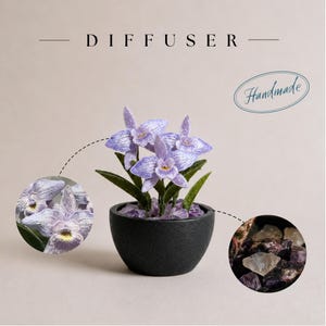 May include: A diffuser featuring a small potted plant with light purple embroidered orchid flowers and green leaves in a matte black bowl. The base is filled with small, translucent, and dark purple stones. The text reads DIFFUSER and Handmade.