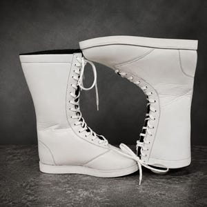 May include: A pair of tall white lace-up boots featuring a smooth leather-like exterior, a high shaft, and white laces threaded through metal eyelets. The interior lining is black, and the boots have a flat, solid white sole construction.