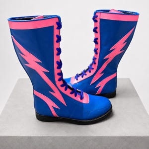 May include: A pair of knee-high boots in bright blue with vibrant pink lightning bolt appliques. The boots feature a lace-up front with dark blue laces, a pink trim along the top collar, and a solid black sole for a bold, high-contrast look.