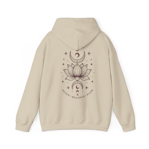 May include: A beige hooded sweatshirt featuring a dark brown graphic on the back. The design displays a central lotus flower flanked by crescent moons and stars, with the text ALIGN BALANCE FLOW printed in a curved line beneath the illustration.