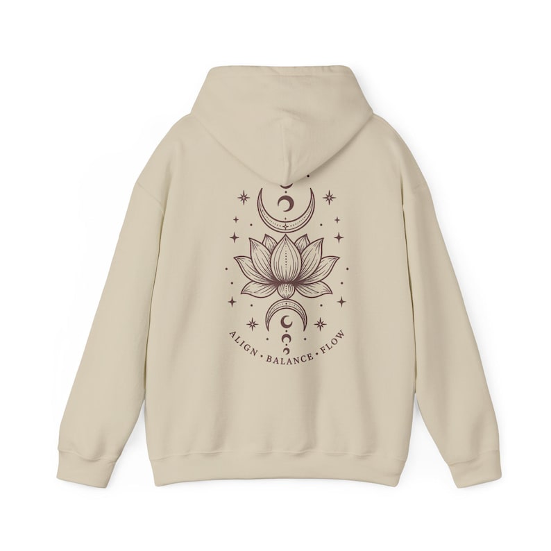 May include: A beige hooded sweatshirt featuring a dark brown graphic on the back. The design displays a central lotus flower flanked by crescent moons and stars, with the text ALIGN BALANCE FLOW printed in a curved line beneath the illustration.