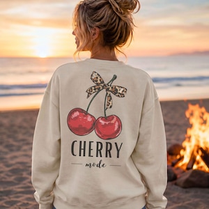 Könnte beinhalten: Ein cremefarbenes Sweatshirt mit Rundhalsausschnitt und einem Grafikdruck auf der Rückseite, der zwei rote Kirschen zeigt, die mit einer Schleife im Leopardenmuster verbunden sind. Unter den Kirschen steht der Text CHERRY mode in dunkler Schrift.