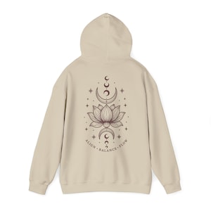 May include: A beige hooded sweatshirt featuring a dark brown graphic on the back. The design displays a central lotus flower surrounded by moon phases and stars, with the text ALIGN BALANCE FLOW printed in a curved line beneath the illustration.