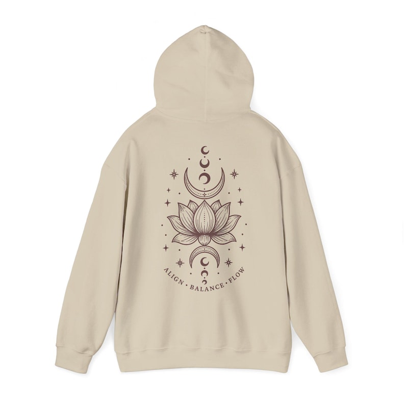 May include: A beige hooded sweatshirt featuring a dark brown graphic on the back. The design displays a central lotus flower surrounded by moon phases and stars, with the text ALIGN BALANCE FLOW printed in a curved line beneath the illustration.