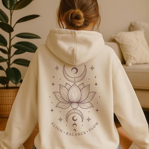May include: A cream-coloured hooded sweatshirt featuring a dark brown graphic on the back. The design includes a central lotus flower, crescent moons above and below, stars, and the text ALIGN, BALANCE, FLOW arranged in a circular pattern.