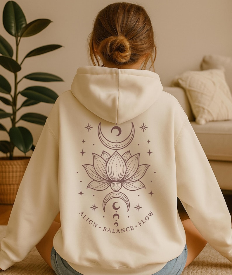 May include: A cream-coloured hooded sweatshirt featuring a dark brown graphic on the back. The design includes a central lotus flower, crescent moons above and below, stars, and the text ALIGN, BALANCE, FLOW arranged in a circular pattern.