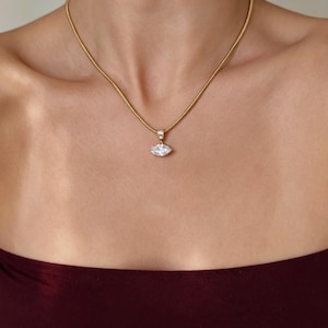 May include: A gold-toned snake chain necklace featuring a single marquise-cut clear gemstone pendant. The pendant is suspended from a small, rectangular bail attached to the chain, which rests against the collarbone in a simple, elegant design.