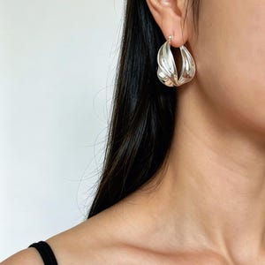 May include: A polished silver hoop earring with a twisted, sculptural design. The earring features a smooth, reflective metallic surface that curves inward, creating a dimensional, teardrop-like shape that hangs from the earlobe.