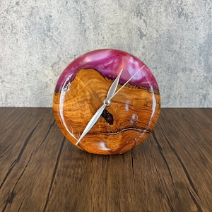 May include: A round clock featuring a natural wood grain center with a polished finish and a vibrant magenta resin border. The clock face includes three thin silver-colored metal hands positioned at the center of the circular wooden piece.