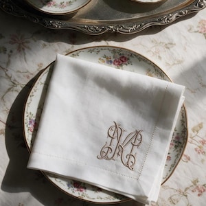 Op de afbeelding: Een vierkant wit stoffen servet met een decoratief, lichtbruin geborduurd monogram van de letters M en K verstrengeld in cursief schrift. Het servet heeft een eenvoudige zoom en ligt netjes gevouwen op een bord met een bloemenpatroon.