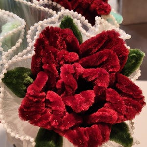 Op de afbeelding: Een handgemaakte bloem van donkerrode, pluizige pijpenragers met donkergroene fluwelen bladeren. De bloem is omringd door een delicate witte kanten rand versierd met kleine witte kraaltjes, wat zorgt voor een textuurrijk en gelaagd bloemstuk.