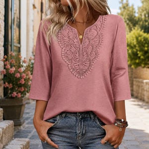 May include: A dusty rose-colored top with three-quarter length sleeves and a V-neckline. The front features an intricate, symmetrical lace panel with scalloped edges that extends from the neckline down to the center of the chest area.