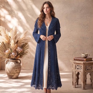 May include: A long, navy blue, open-front knit duster cardigan featuring long sleeves, a textured cable knit pattern, and a fringed shawl collar. The garment is worn over a simple, floor-length, cream-colored sleeveless dress with a scoop neckline.
