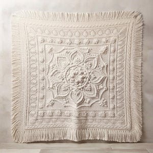 Puede incluir: Una manta cuadrada de color blanco roto con un intrincado patrón central de mandala con texturas en relieve de punto de cable, bodoques y puntos popcorn. La manta está enmarcada por un borde decorativo y terminada con largos flecos.